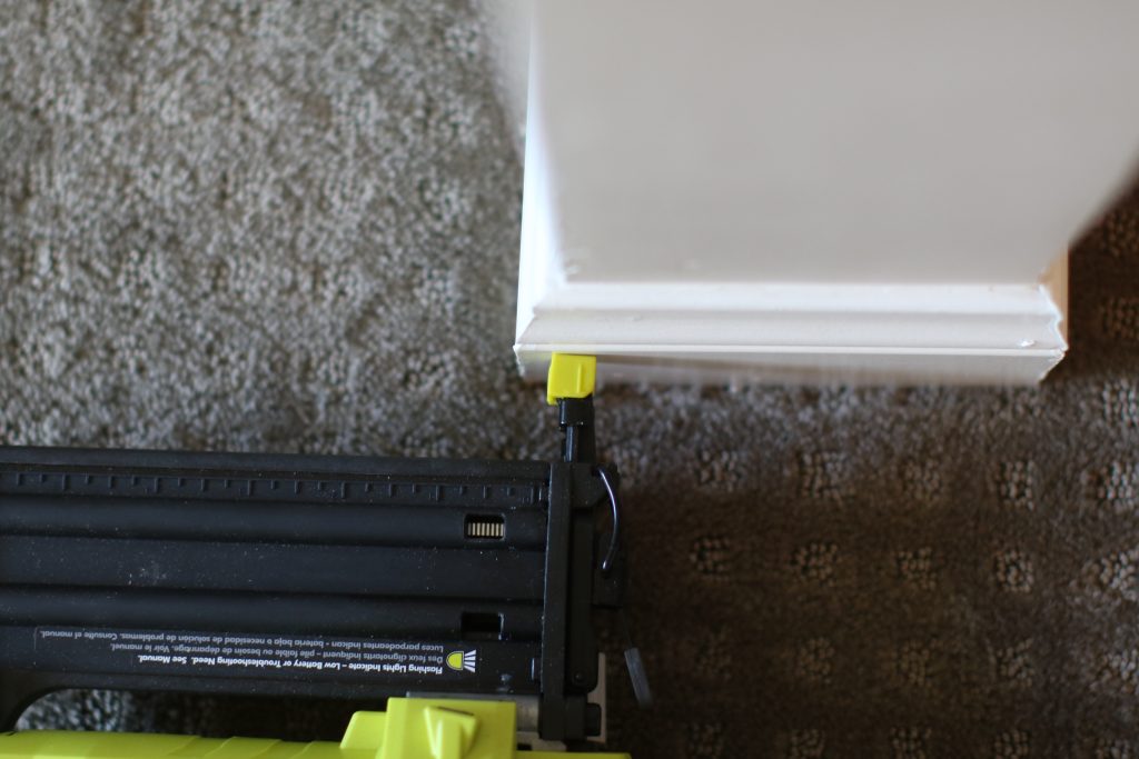 A green and black nailer is being used on a white wall near the gray carpet floor