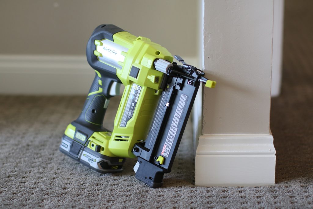 A green and black colored nailer was placed against the white wall on top of a gray carpet