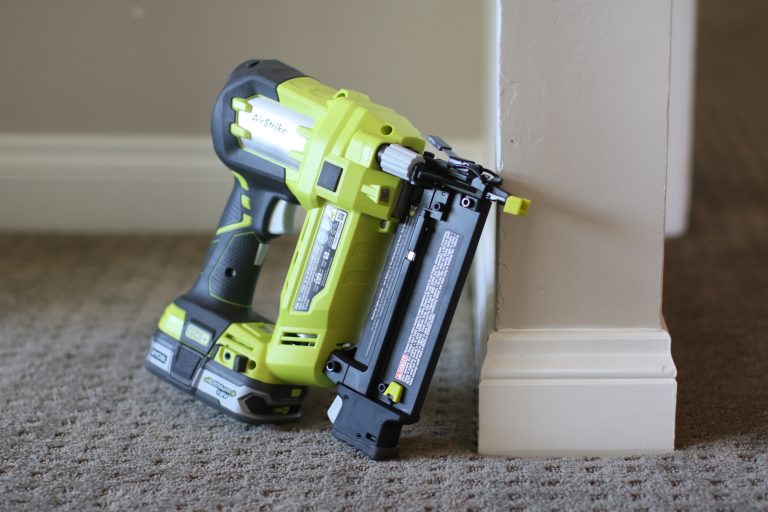 A green and black colored nailer was placed against the white wall on top of a gray carpet