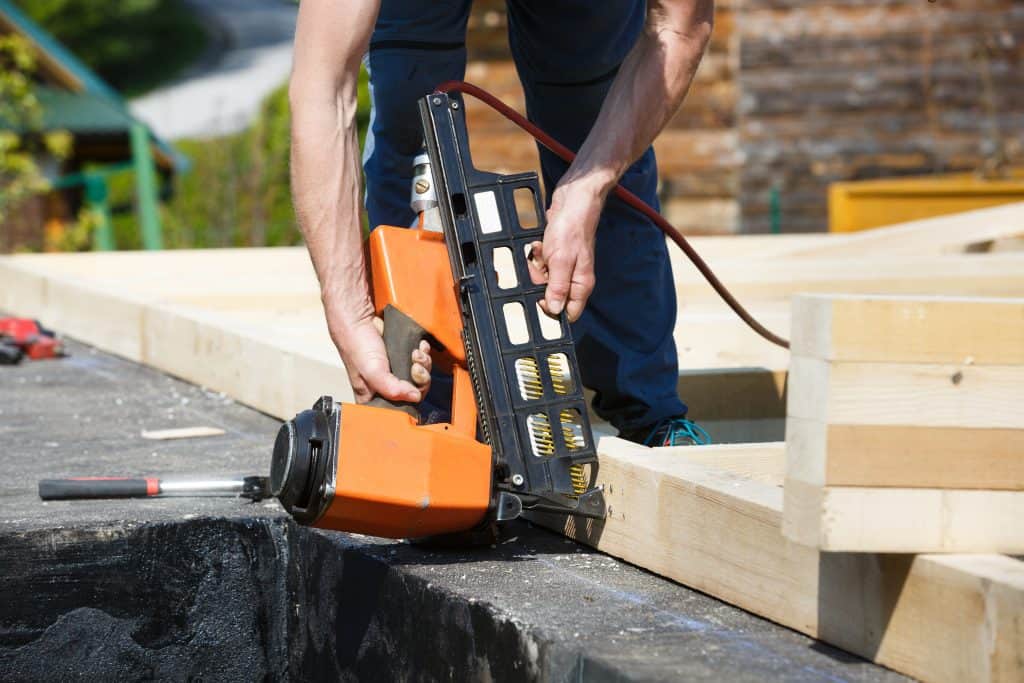 A person wearing blue pants is using an orange and black colored nailer with gold colored nails on a wood