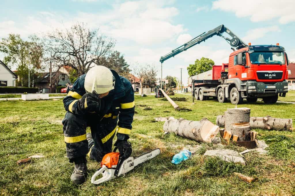 A man wearing blue and yellow coveralls is holding an orange and white colored chainsaw placed on the green grass