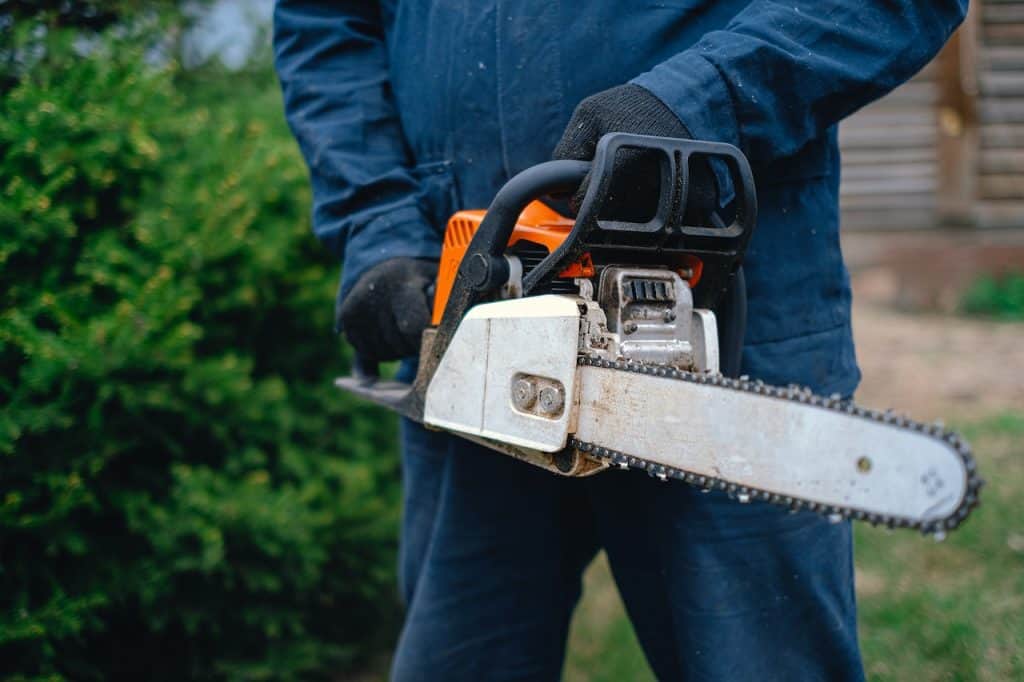 A man wearing a blue coveralls is holding an orange and black colored chainsaw with a silver colored blade near the bush