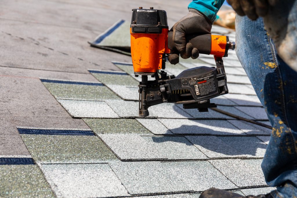 A person wearing denim pants is using a black and orange roofing nailer on gray roof shingles