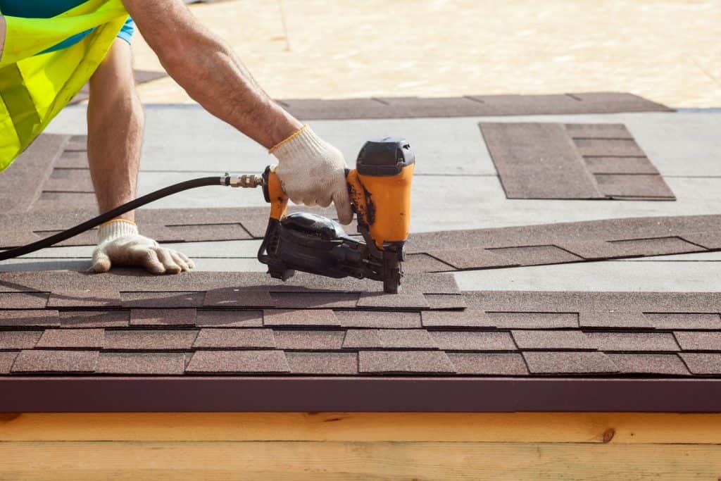 A person wearing a yellow reflective vest and white gloves is holding an orange and black roof nailer on a roof