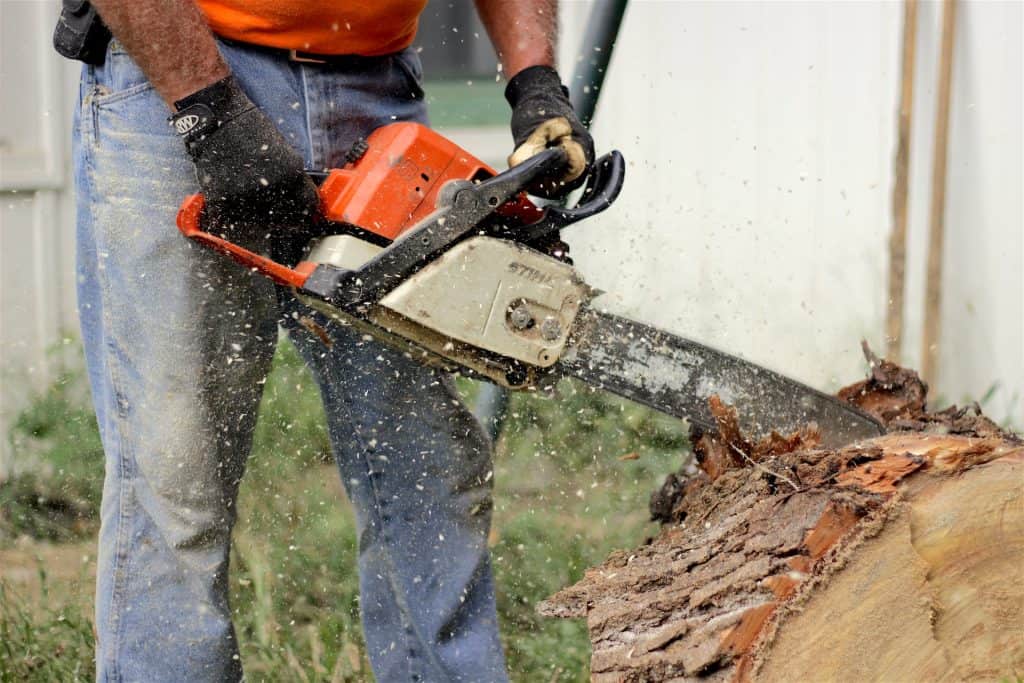 A man wearing black gloves is holding a gray and orange chainsaw to cut a brown chunk of wood