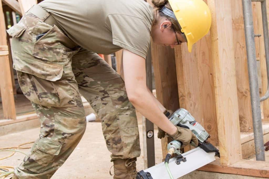 Woman in camouflage uniform and yellow protective hat using a framing nailer to fasten the foundation of wooden pallets
